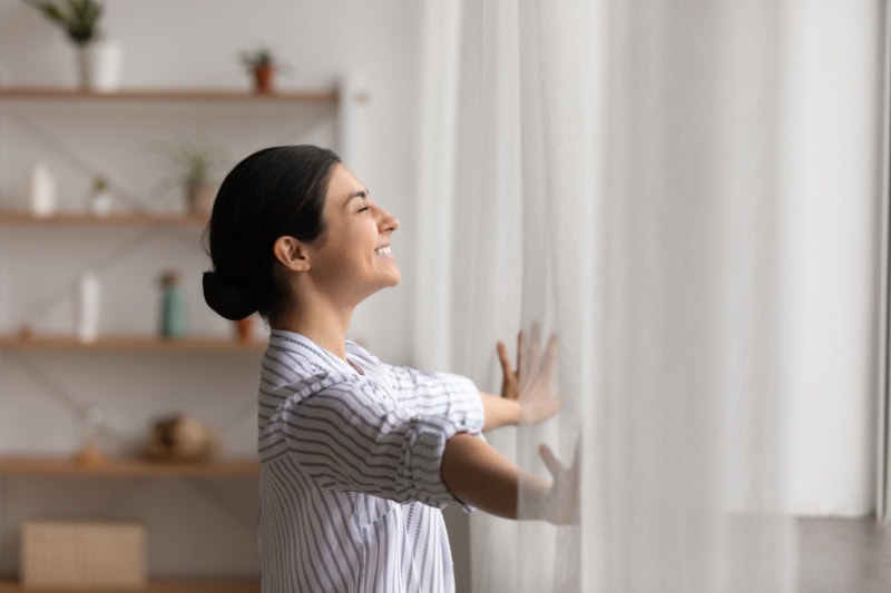 Is It Worth Installing a Ductless Air Conditioner? Bathing in sunlight. Excited young indian lady meet first morning at new flat house part curtains enjoy being homeowner. Happy hindu female open drapes on window breath fresh air close eyes in delight.